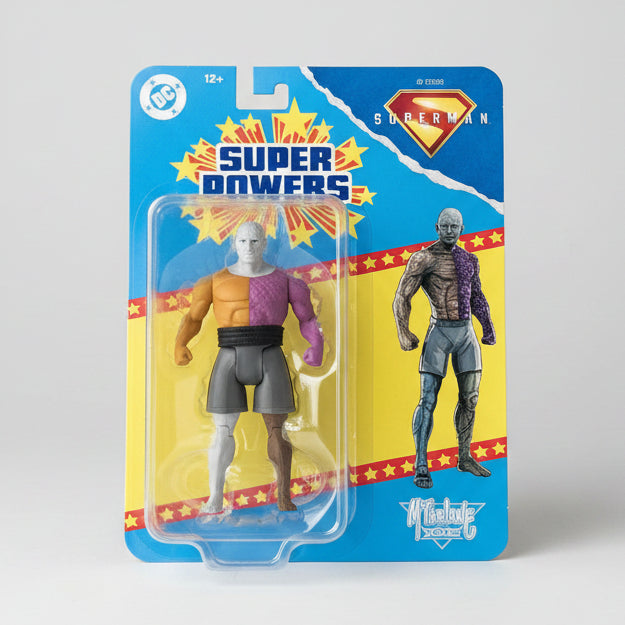 A DC Comics Superman action figure in its original blister card packaging, featuring the character with a white head, purple and orange body, and gray shorts, alongside a larger printed figure on the card.